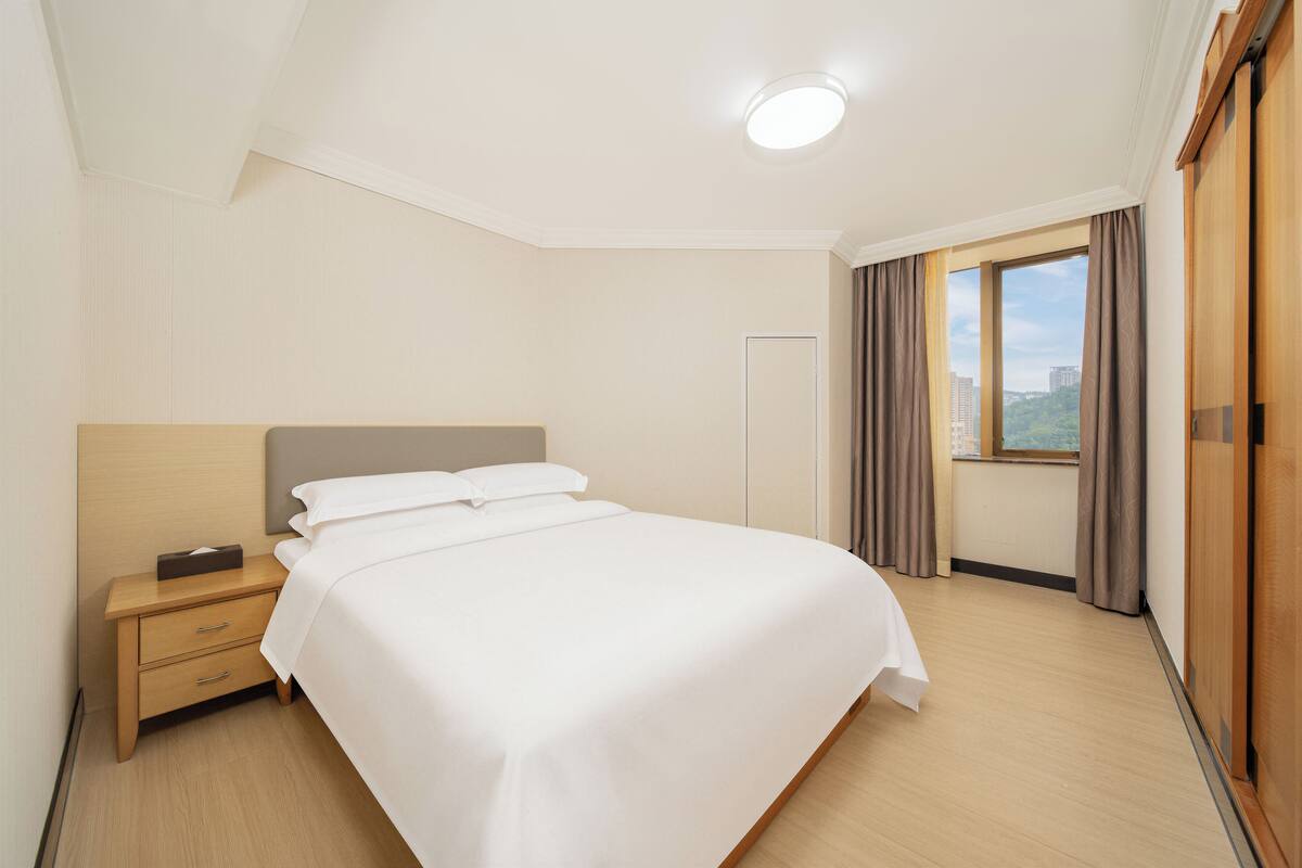 Ein Hotelzimmer mit Bett, Nachttisch, Fenster mit Vorhängen und Blick auf die Stadt.
