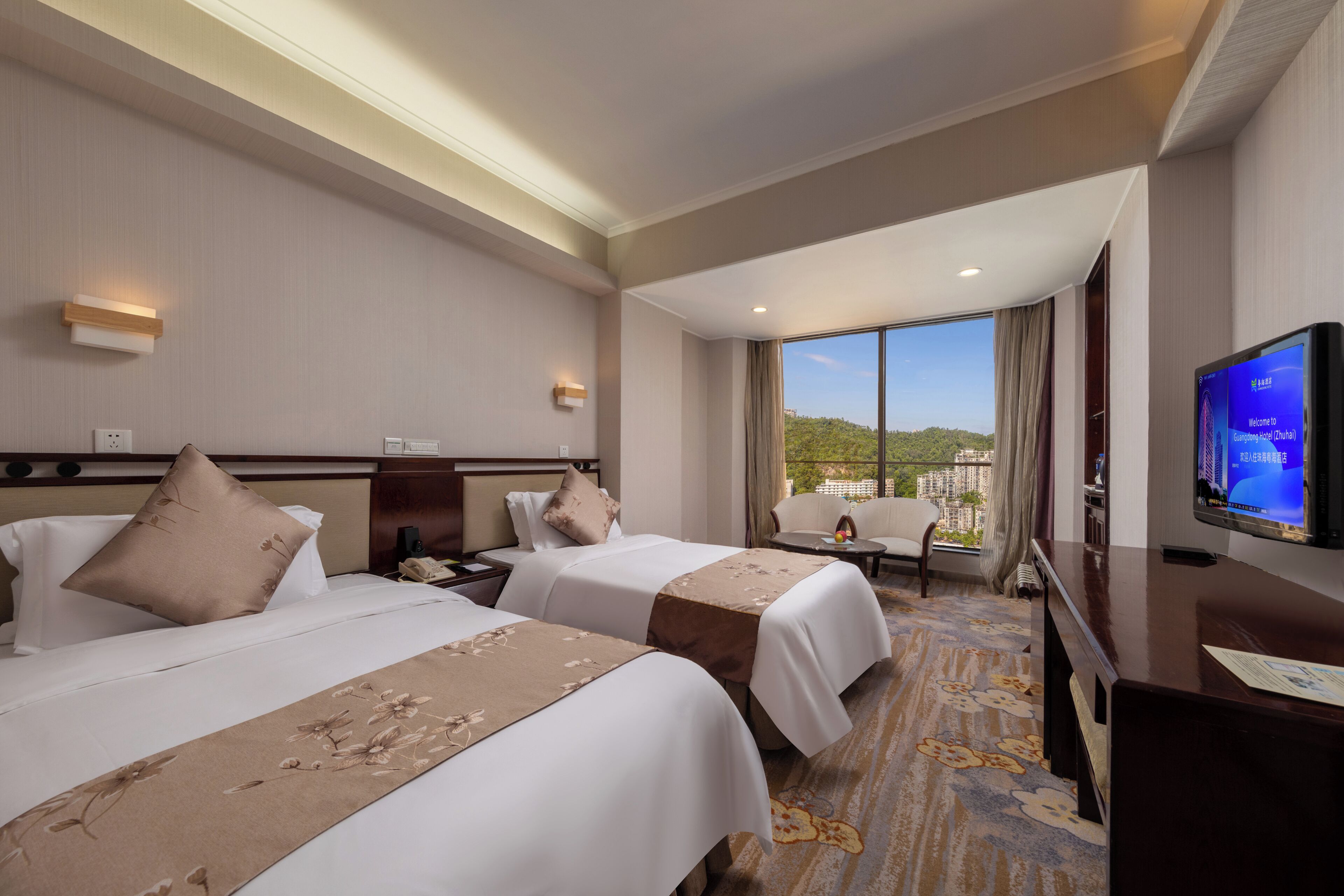 A hotel room with two beds, a TV, a desk, and a large window with a view of a cityscape.