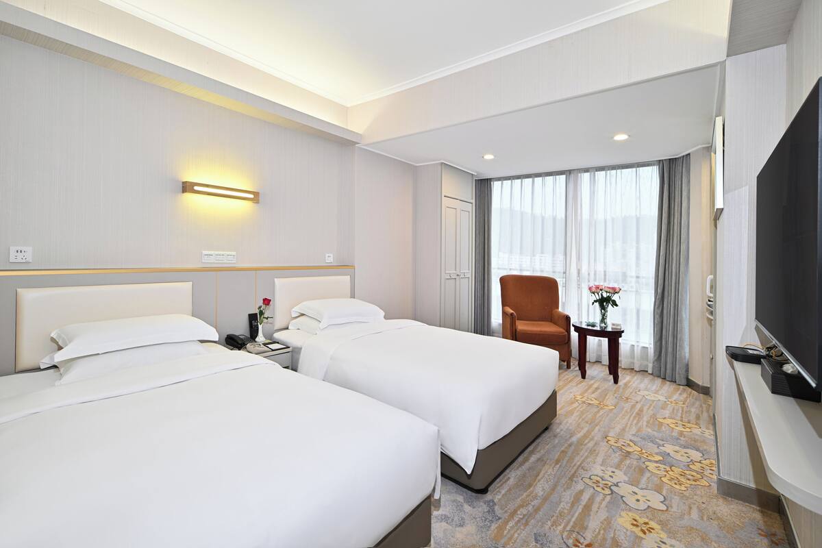 Ein Hotelzimmer mit zwei Betten, einem Fernseher, einem Sessel, einem kleinen Tisch mit einer Vase und einem großen Fenster mit Vorhängen.