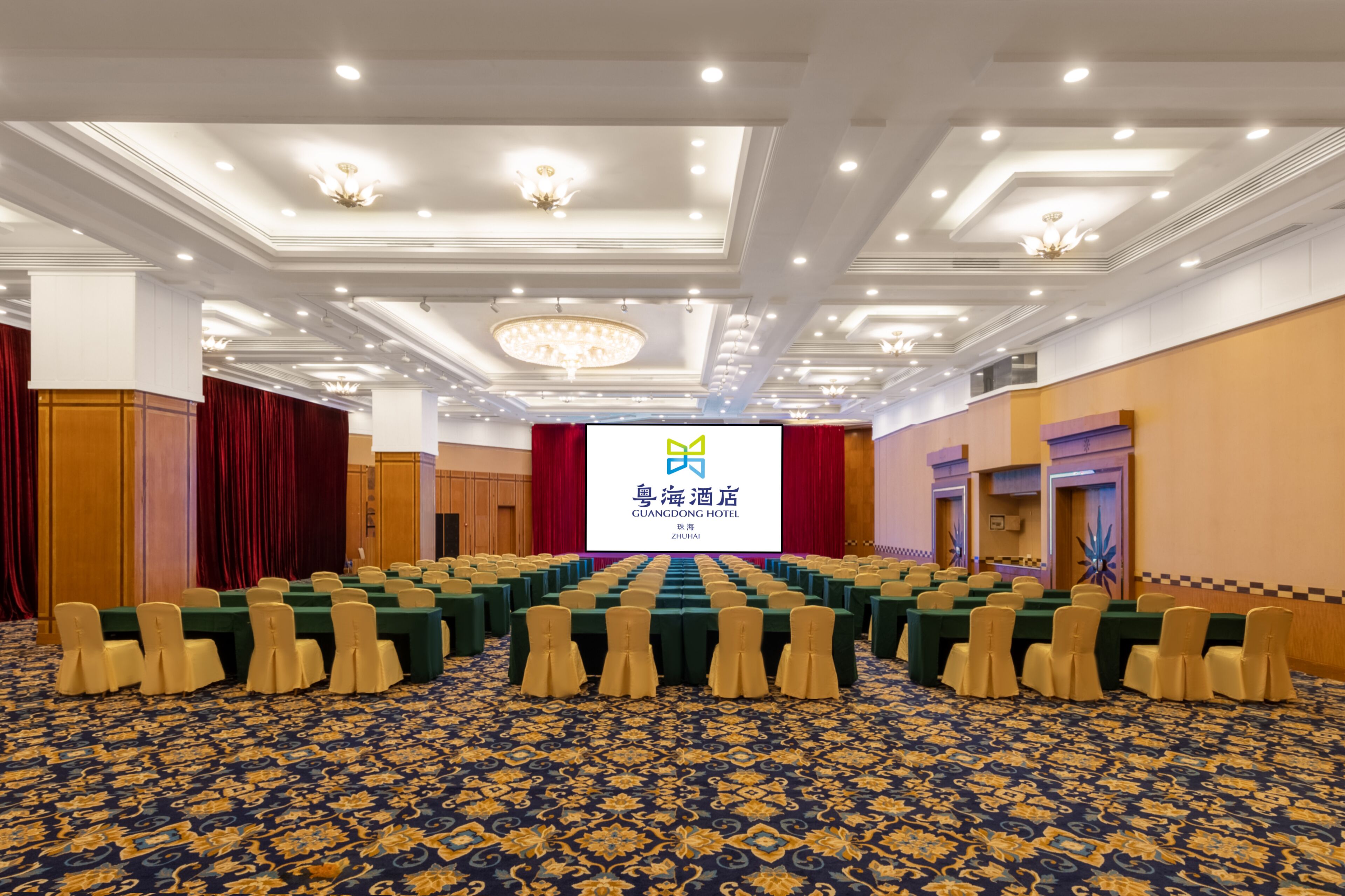 A conference room with rows of tables and chairs, a large screen displaying hotel information, and a patterned carpet.