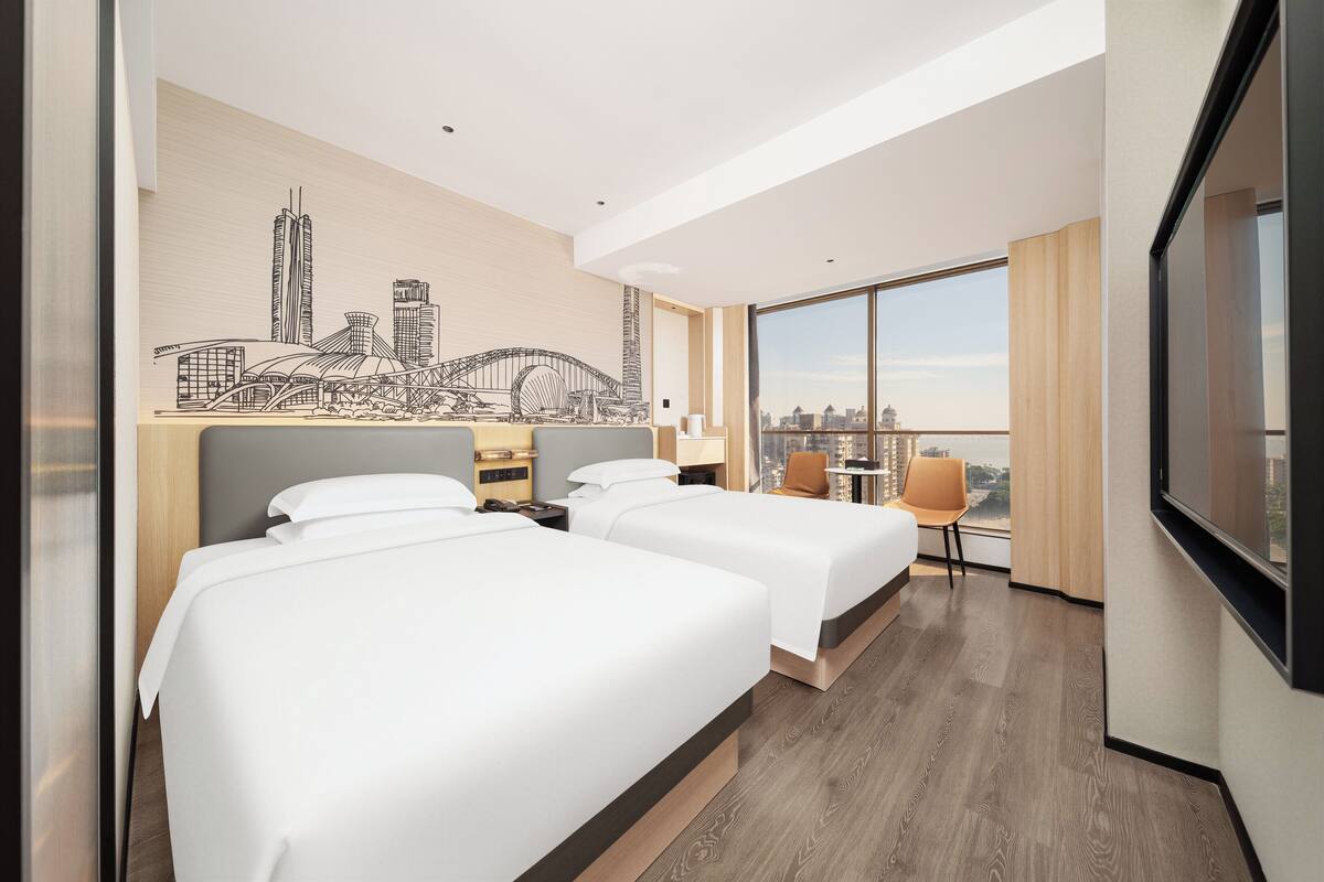 Ein Hotelzimmer mit zwei Betten, einer Stadt-Mural, einem Essbereich und einem großen Fenster mit Blick auf die Stadt.