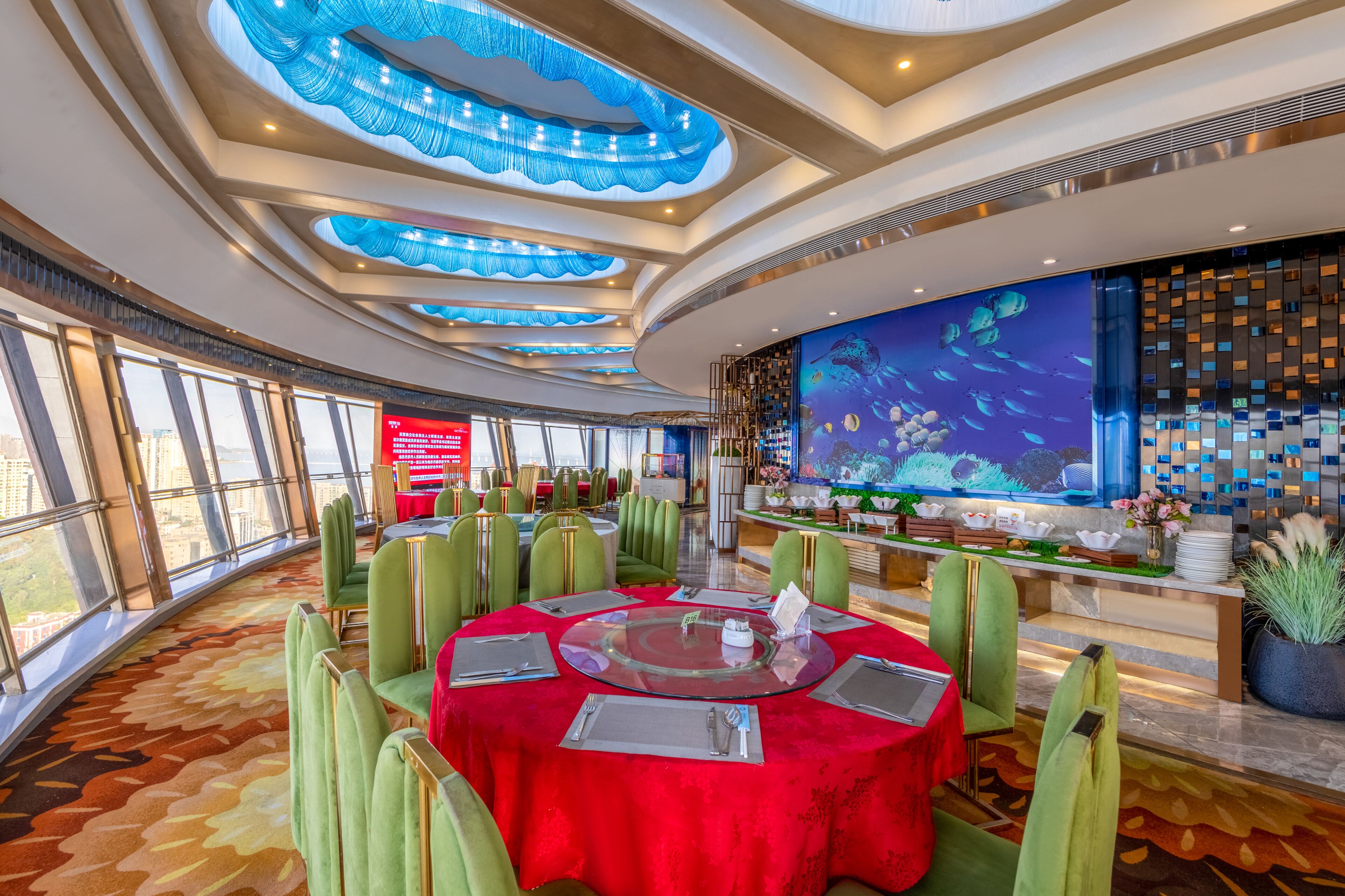 A spacious dining area with round tables, green upholstered chairs, and a large aquarium display on the wall.