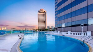 A swimming pool with a curved edge, surrounded by a tiled deck and a white railing. In the background, there is a tall building with a curved top and a modern glass facade. - Guangdong Hotel Zhuhai (Zhuhai)