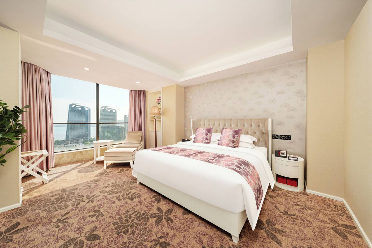 Ein Hotelzimmer mit einem großen Bett, einem Nachttisch, einem Sessel und Blick auf die Stadt.