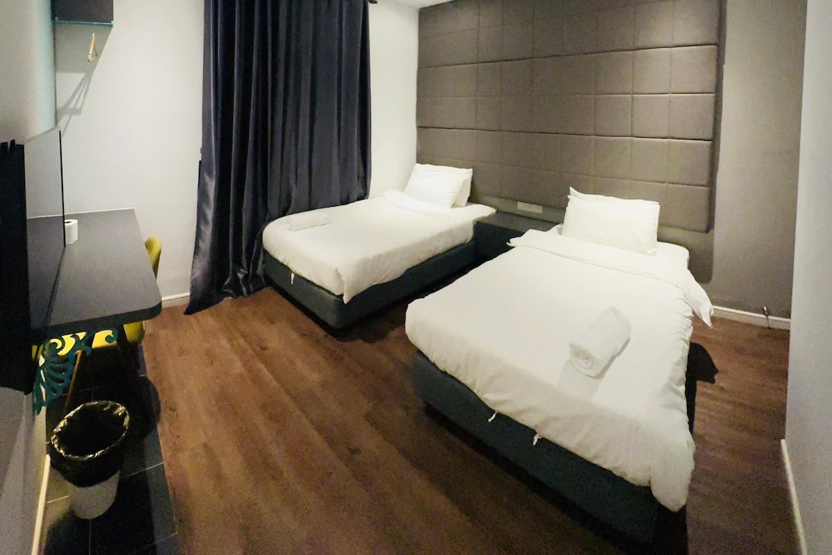A hotel room with two beds, a desk, a chair, and a trash bin.