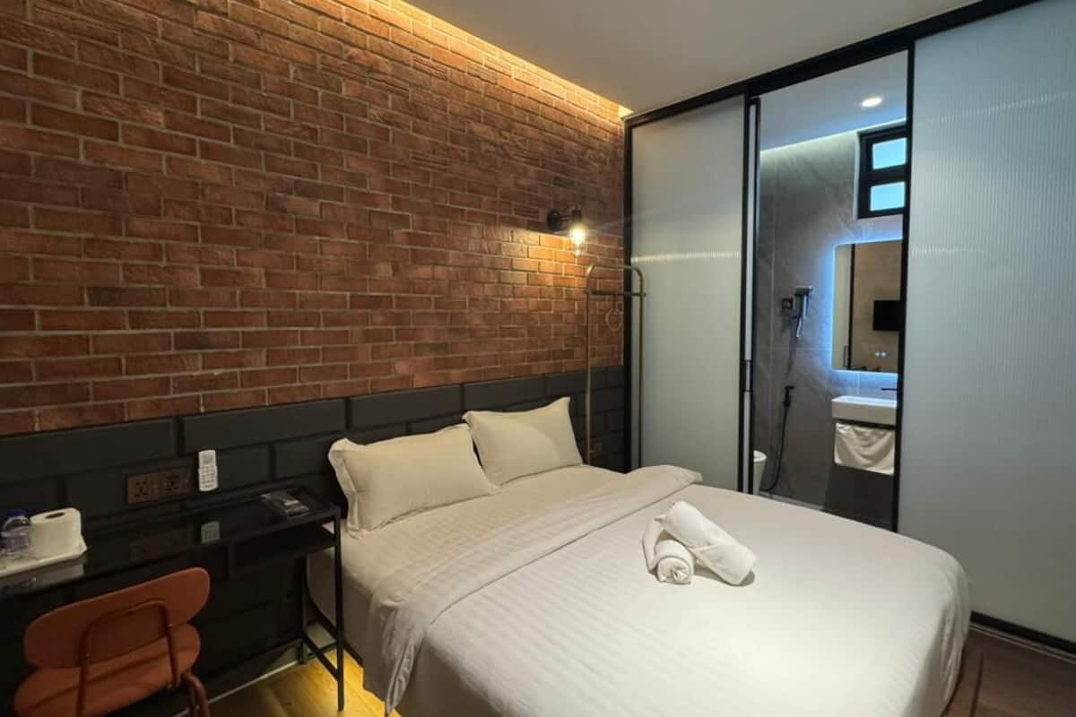 A hotel room with a brick wall, a bed with white linens, a desk with a chair, and a sliding door leading to a bathroom.
