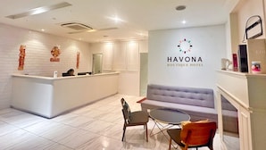 Ein Hotel-Rezeptionsbereich mit einem weißen Empfangstresen, einer Couch und einem kleinen Tisch. Der Hotelname "HAVONA BOUTIQUE HOTEL" ist an der Wand sichtbar.