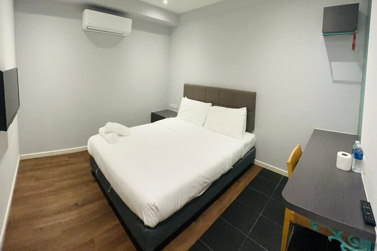 A hotel room with a bed, a desk, a chair, a TV, and an air conditioning unit.