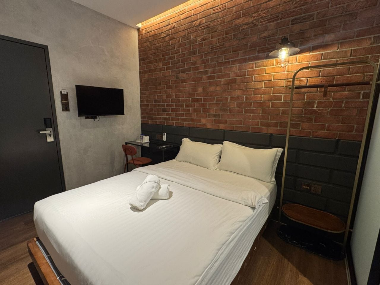 A hotel room with a brick-patterned headboard, a flat-screen TV mounted on the wall, a bedside table with a lamp, and a neatly made bed with a towel.