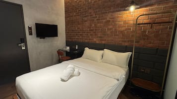 A hotel room with a brick-patterned headboard, a flat-screen TV mounted on the wall, a bedside table with a lamp, and a neatly made bed with a towel.