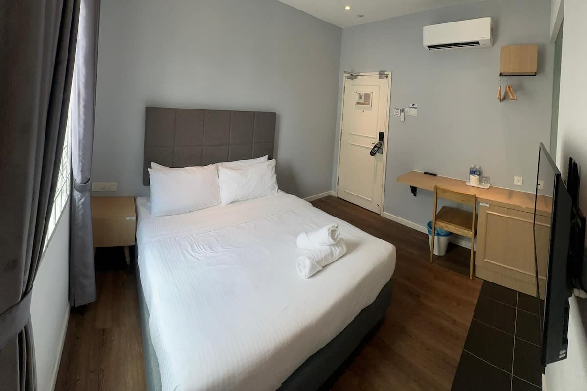A hotel room with a large bed, a desk, a chair, an air conditioner, and a TV.