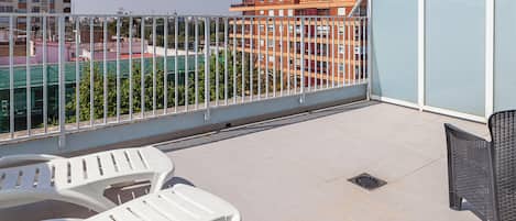 A rooftop terrace with white plastic chairs and a black wicker chair, a metal railing, and a view of surrounding buildings.