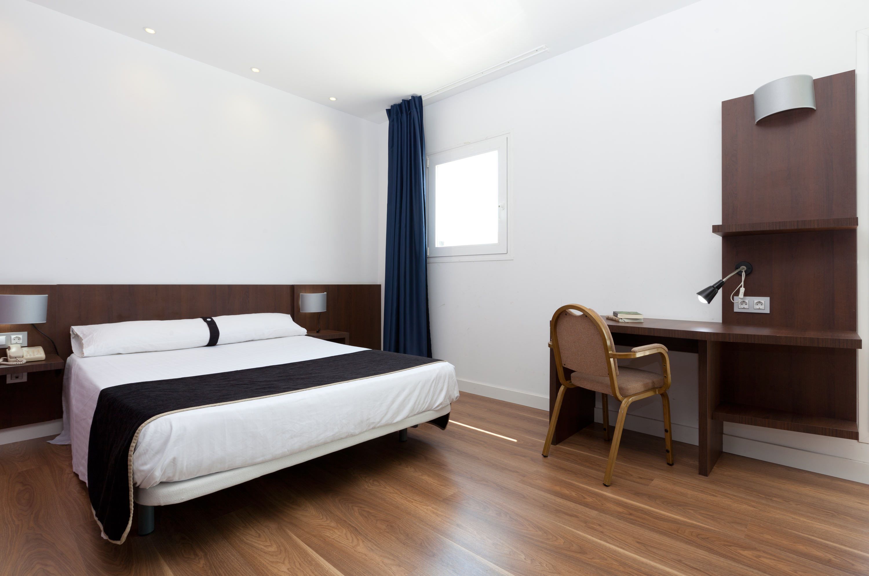 Habitación de hotel con cama, escritorio, silla, lámpara, teléfono y ventana con cortinas.