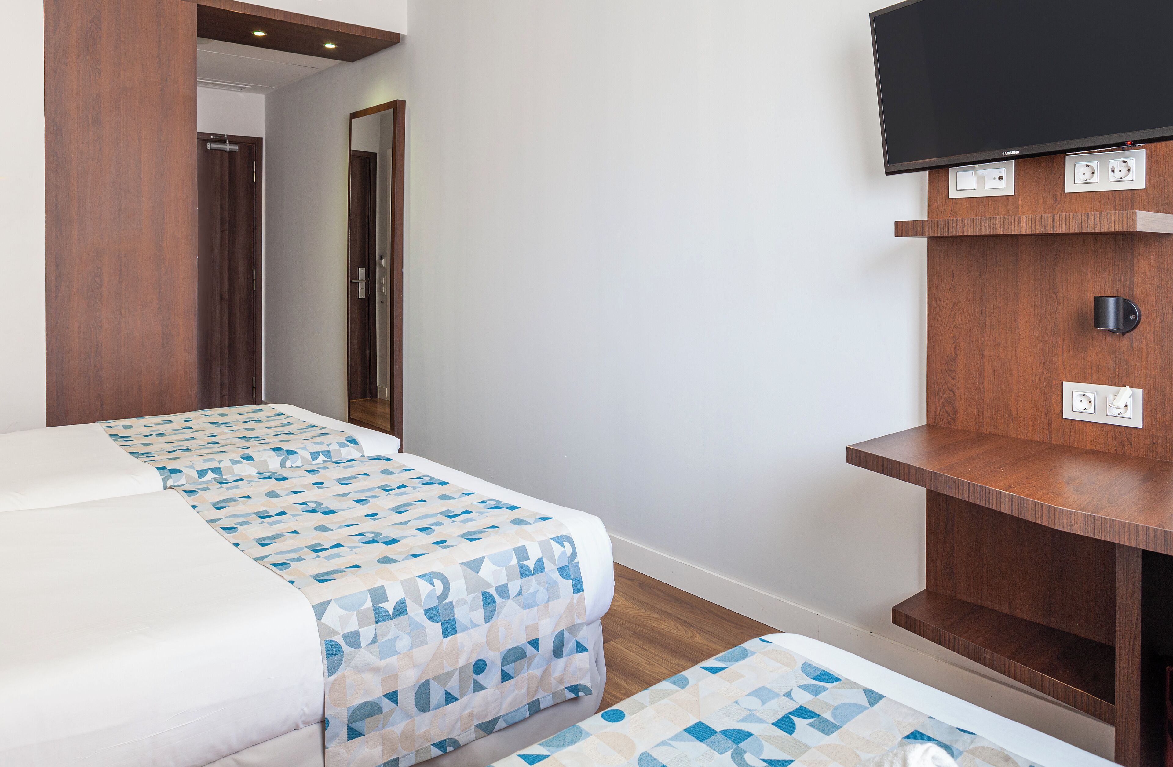 Una habitación de hotel con una cama, un televisor montado en un estante de madera y una puerta que da a otra habitación.