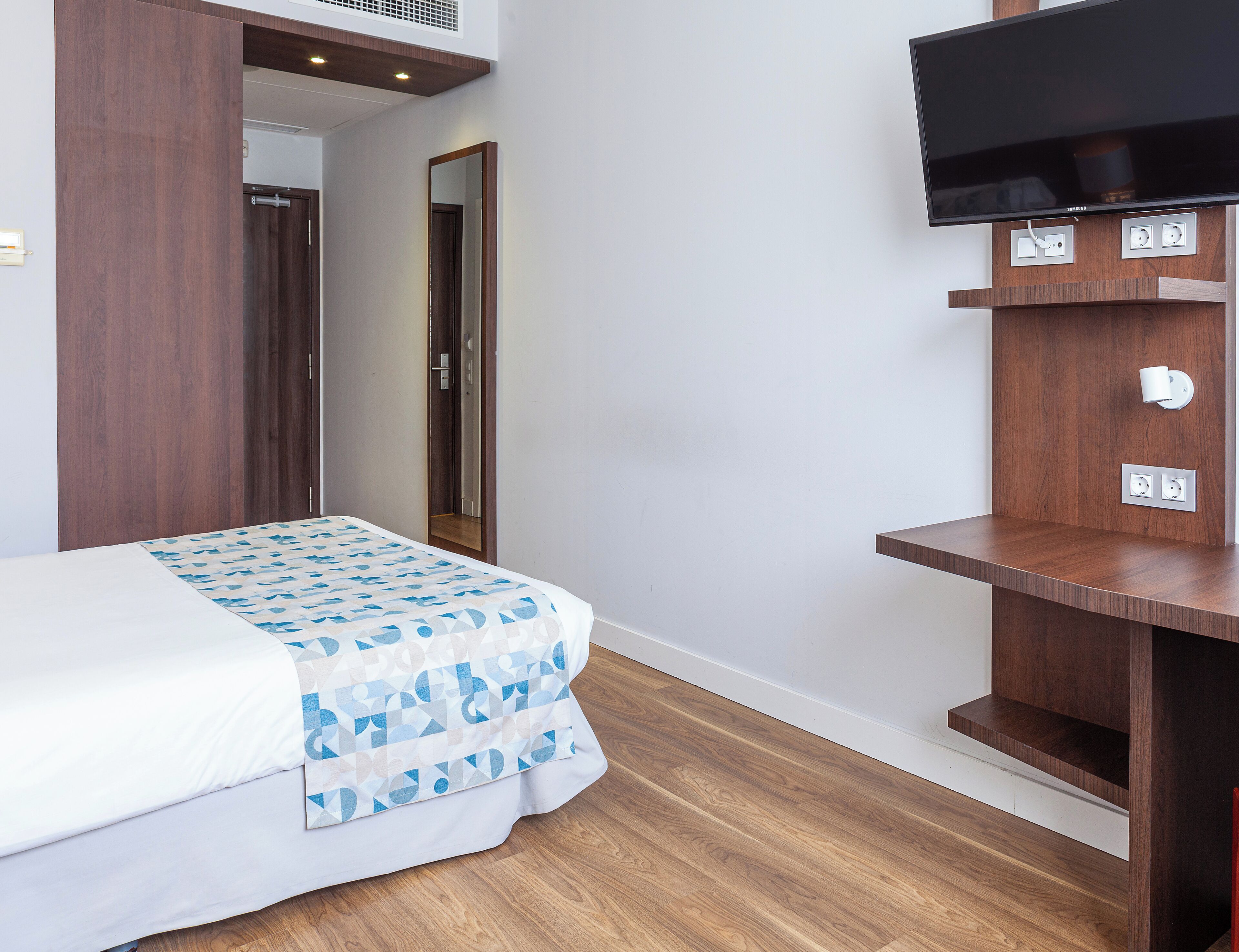 A bedroom with a bed, a TV mounted on a wooden shelf, and a door leading to another room.