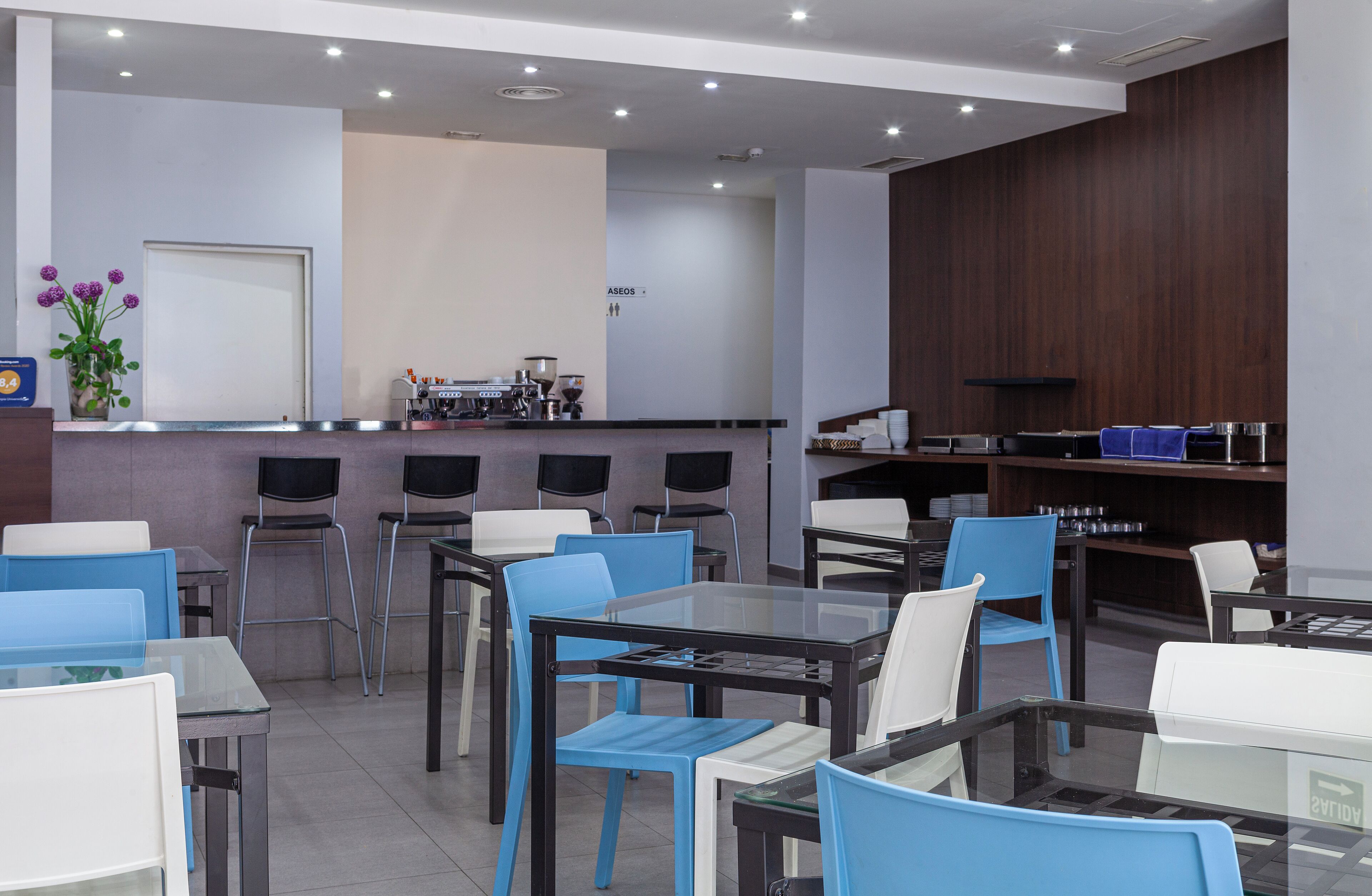 Un cafè modern amb taules i cadires de vidre, una màquina de cafè i una barra amb un menú.