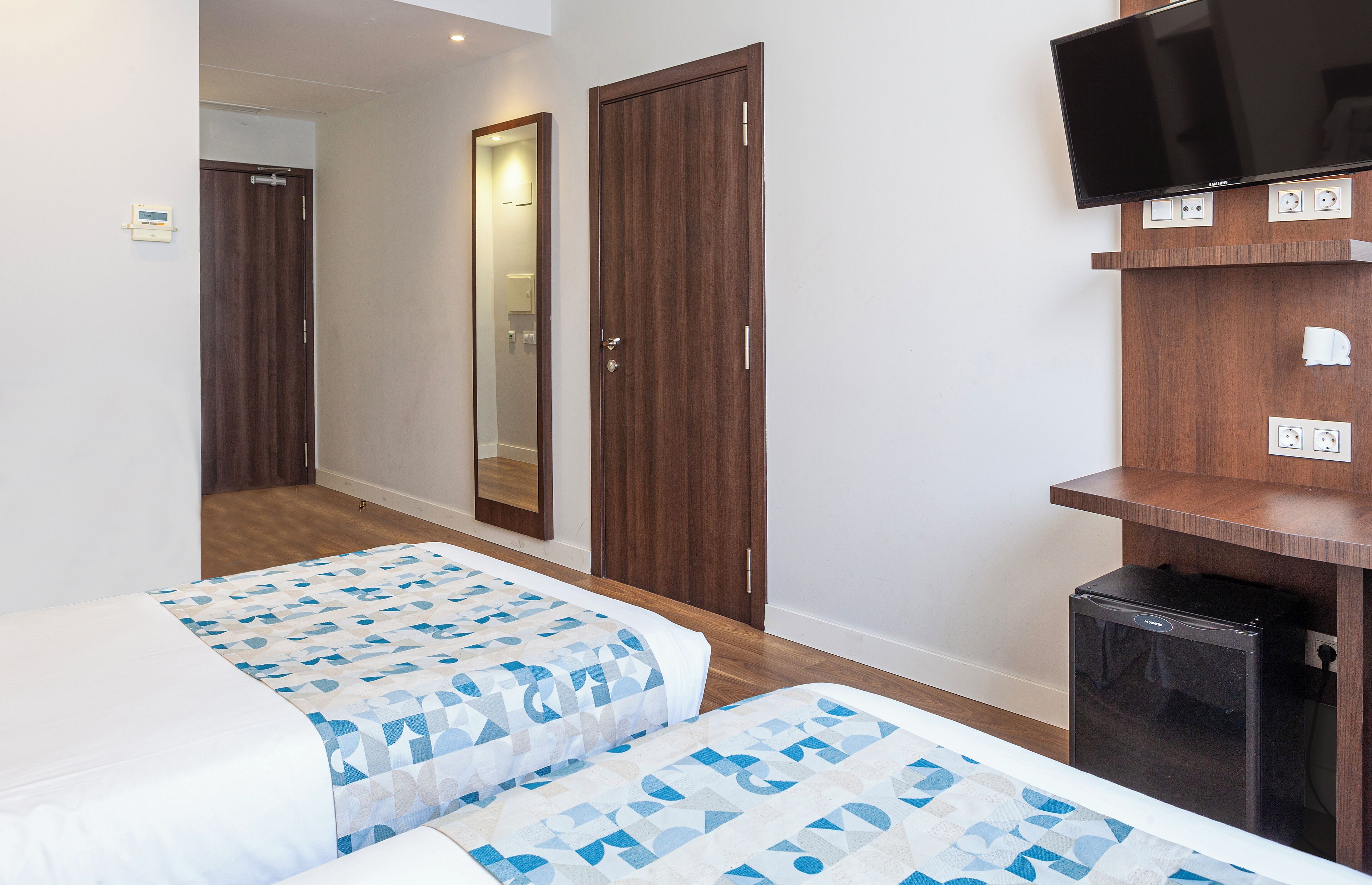 A hotel room with two beds, a TV, a small fridge, and a mirror.