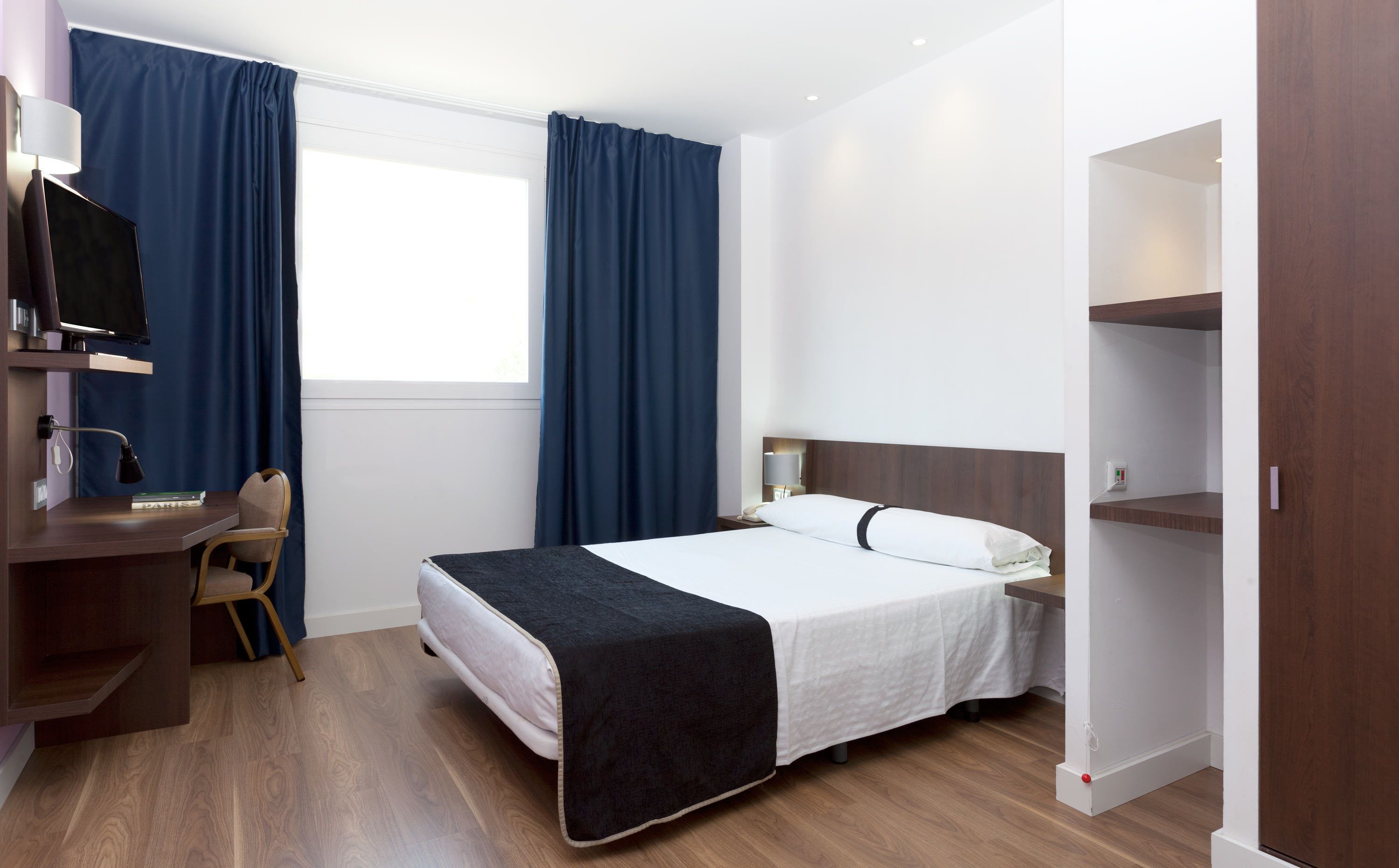 Habitación de hotel con cama, escritorio con silla, televisor y ventana con cortinas.