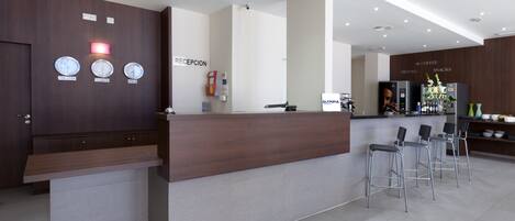 A modern reception area with a wooden reception desk, a clock displaying New York, Valencia, and Tokyo times, and a coffee bar with seating.