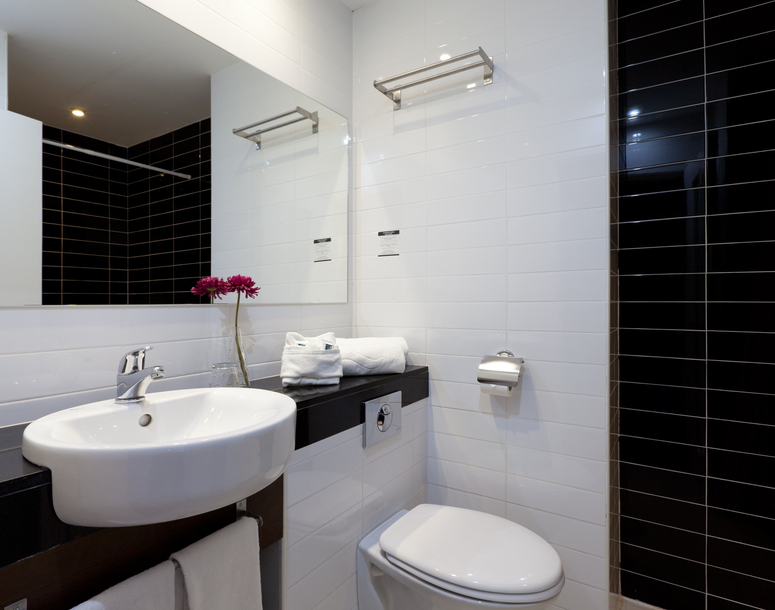 A modern bathroom with a white sink, black countertop, and a white toilet. There is a mirror above the sink and a towel rack on the wall.
