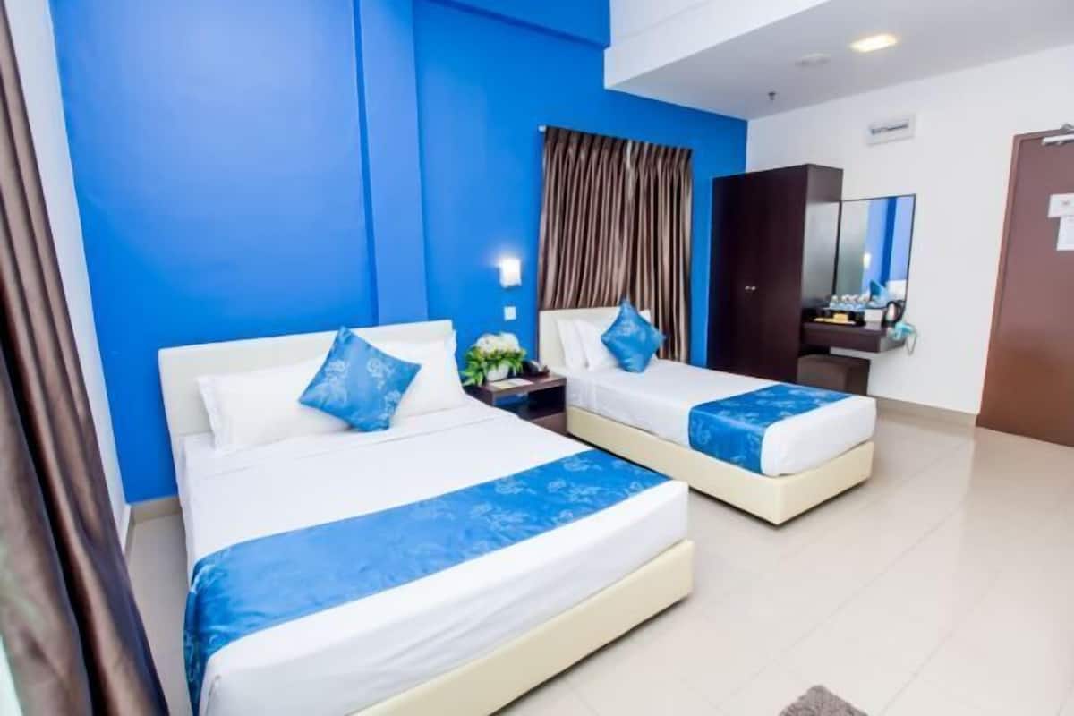 Habitación de hotel con dos camas, cada una con cubrecama azul y almohadas blancas. Hay una mesita de noche con una lámpara y una pequeña mesa con una planta. La habitación tiene una pared azul decorativa y un espejo grande sobre la cama.