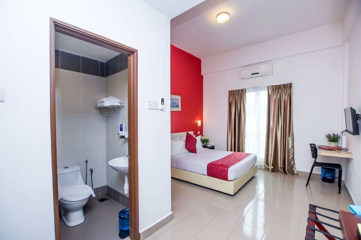 Una habitación de hotel con cama, mesita de noche, escritorio, silla, ventana con cortinas, y baño con inodoro, lavabo y ducha.