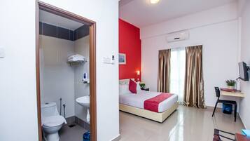 Una habitación de hotel con cama, mesita de noche, escritorio, silla, ventana con cortinas, y baño con inodoro, lavabo y ducha.