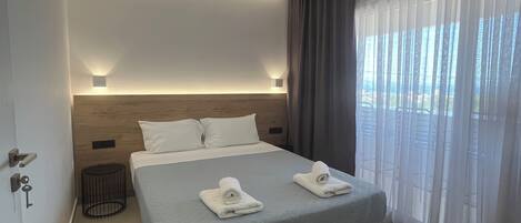 Une chambre d’hôtel moderne avec un grand lit, une table de chevet, une lampe et une vue sur l’extérieur à travers des rideaux légers.