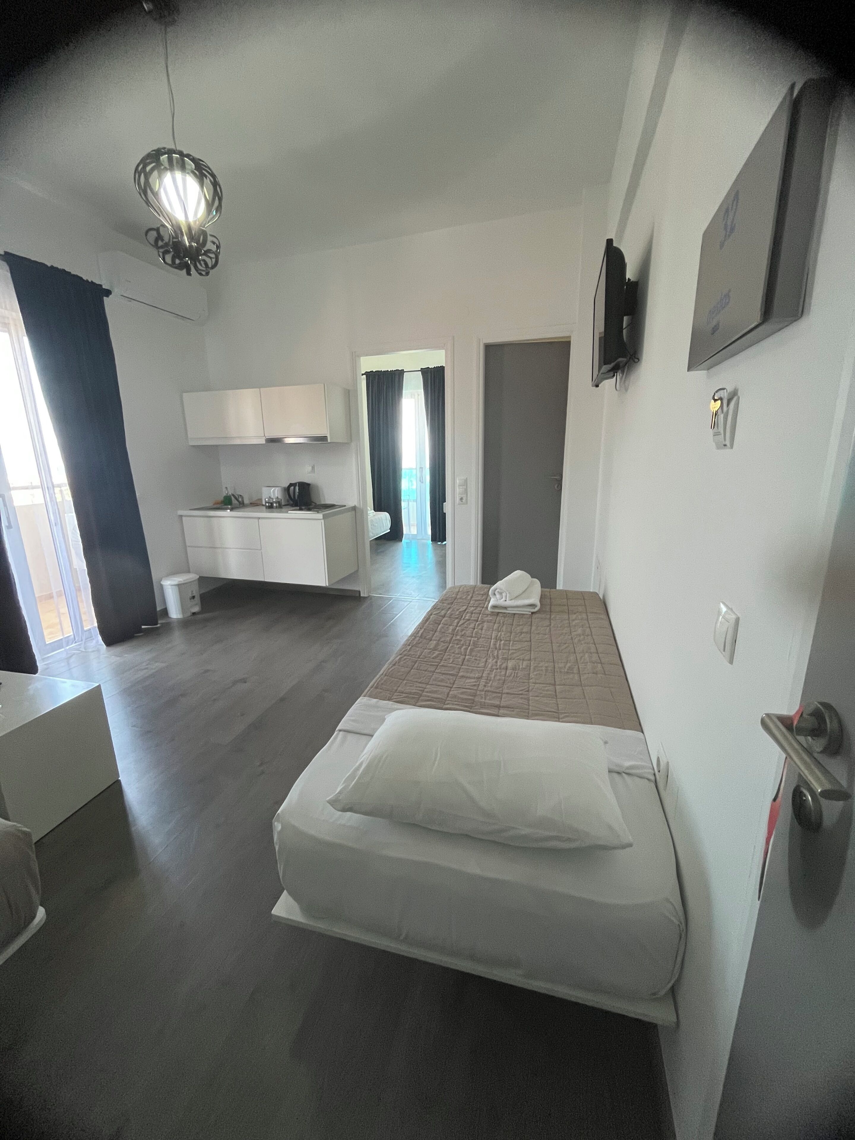 Un dormitorio con cama, mesita de noche, armario, zona de cocina con armarios, lámpara colgante y una puerta que da a otra habitación.