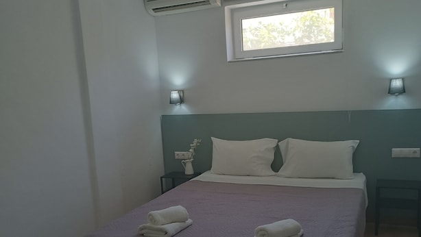 A bedroom with a bed, two bedside tables, a window, and an air conditioning unit. - Aestas Apts (Chania)
