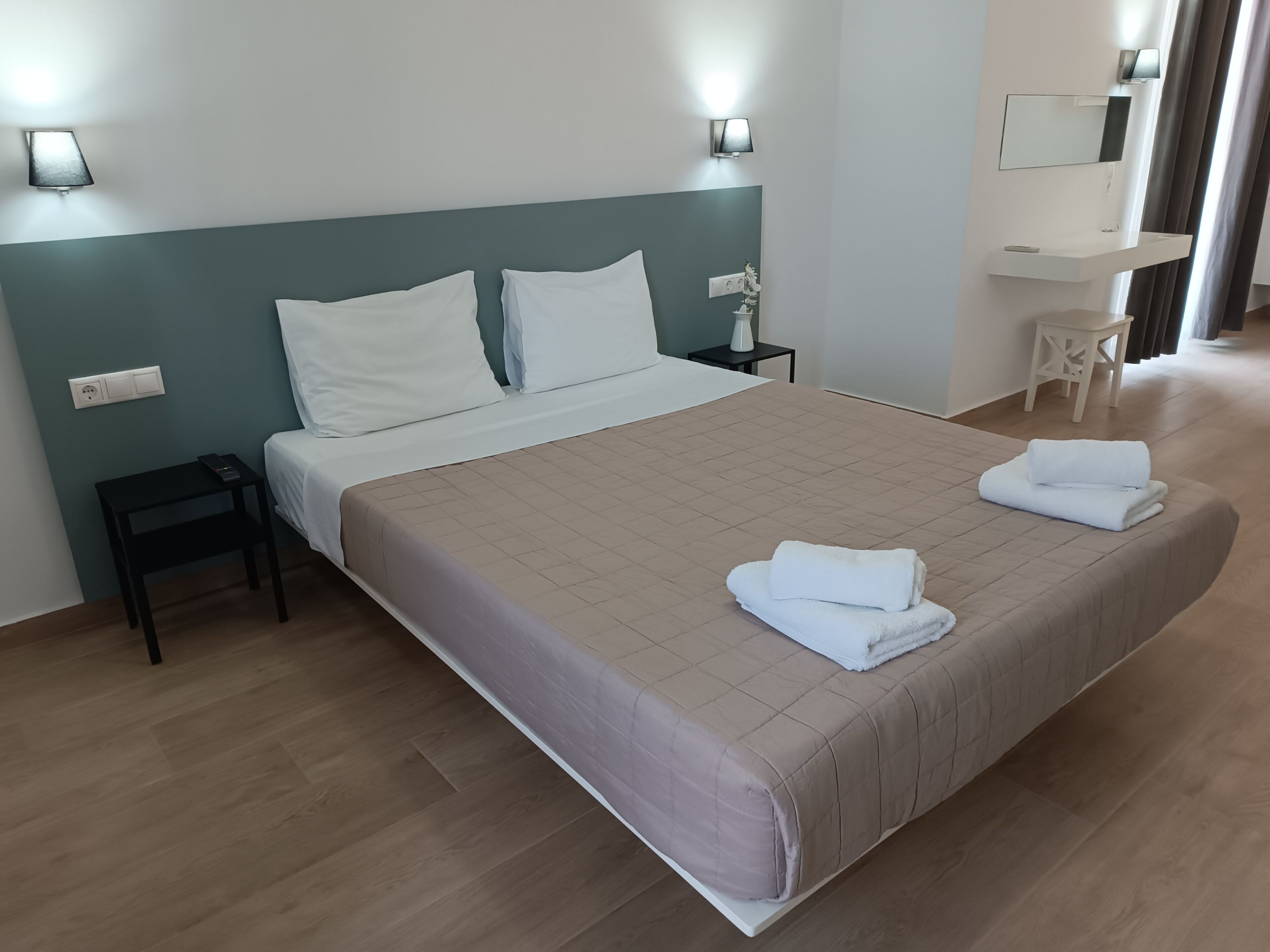 A neatly made bed with a beige bedspread, two white pillows, and two folded towels on the bed. There is a small bedside table with a lamp, a power outlet, and a wall-mounted light fixture.