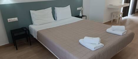 A neatly made bed with a beige bedspread, two white pillows, and two folded towels on the bed. There is a small bedside table with a lamp, a power outlet, and a wall-mounted light fixture.