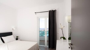 A hotel room with a bed, a small bedside table, a potted plant, a sliding glass door to a balcony, and a mirror on the wall. - Aestas Apts (Chania)