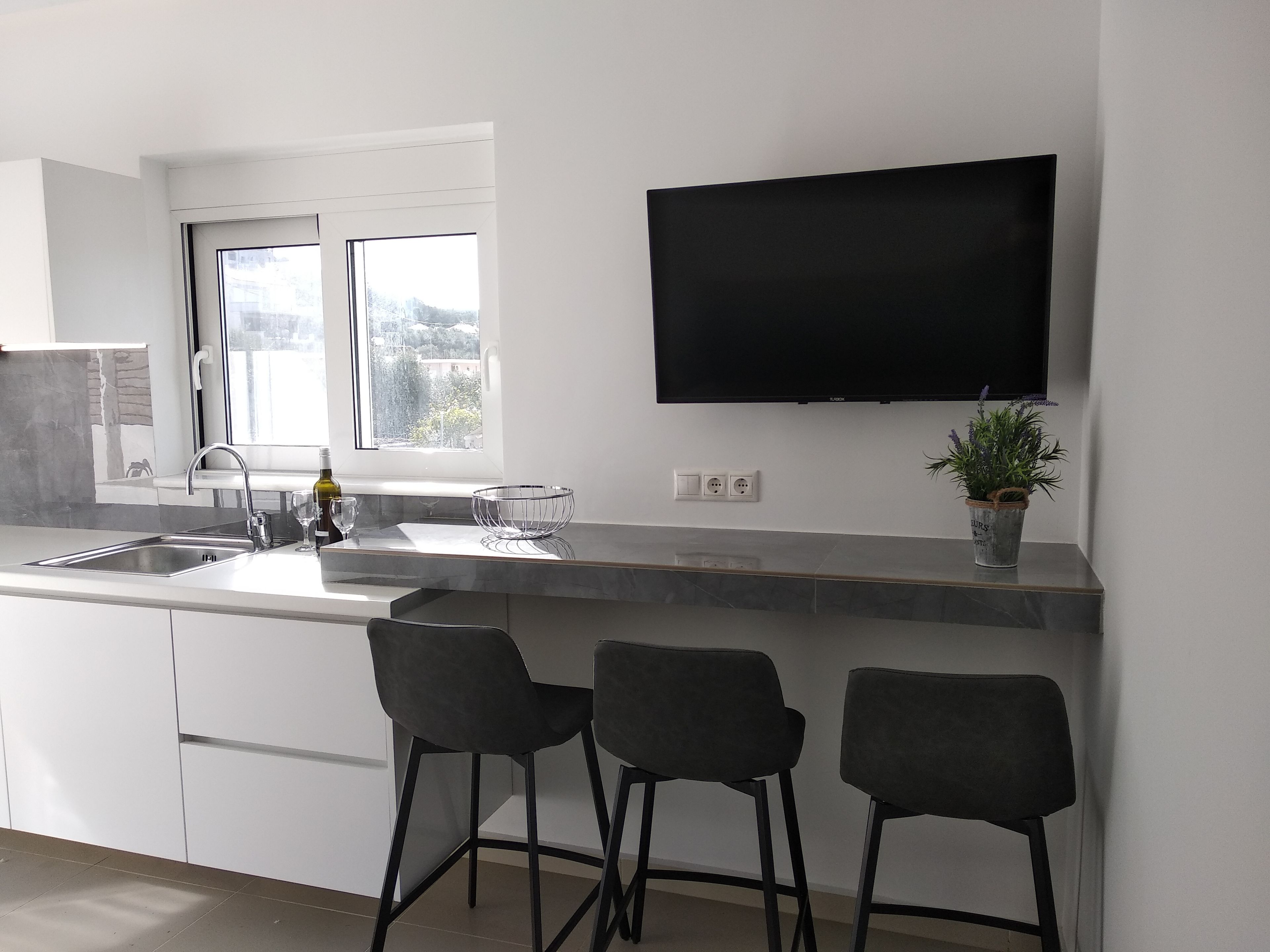 A modern kitchen with a large window, a flat-screen TV, and three black bar stools.