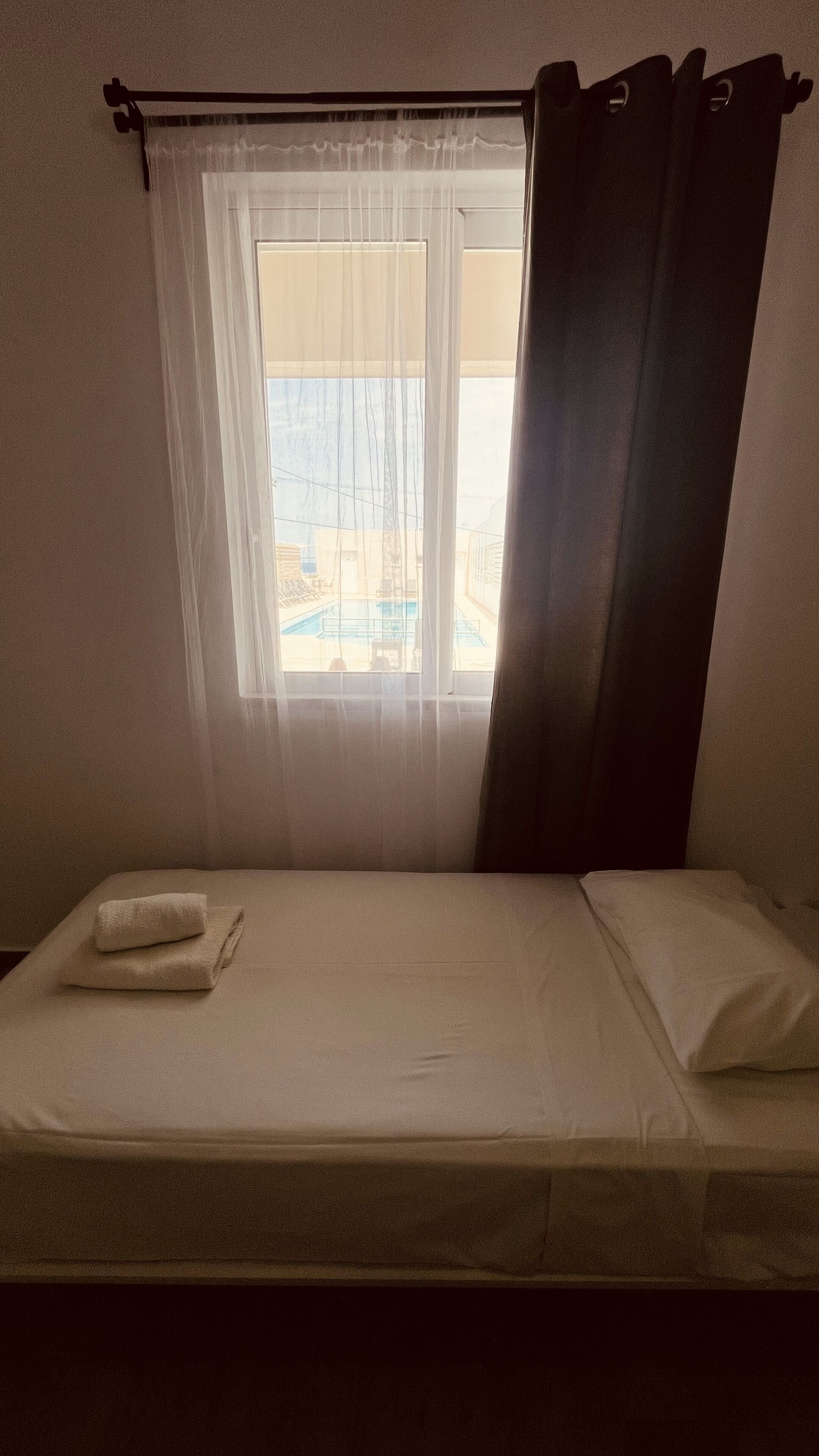 A single bed with a white mattress and a folded white towel, a window with sheer curtains, and a view of buildings and a pool outside.