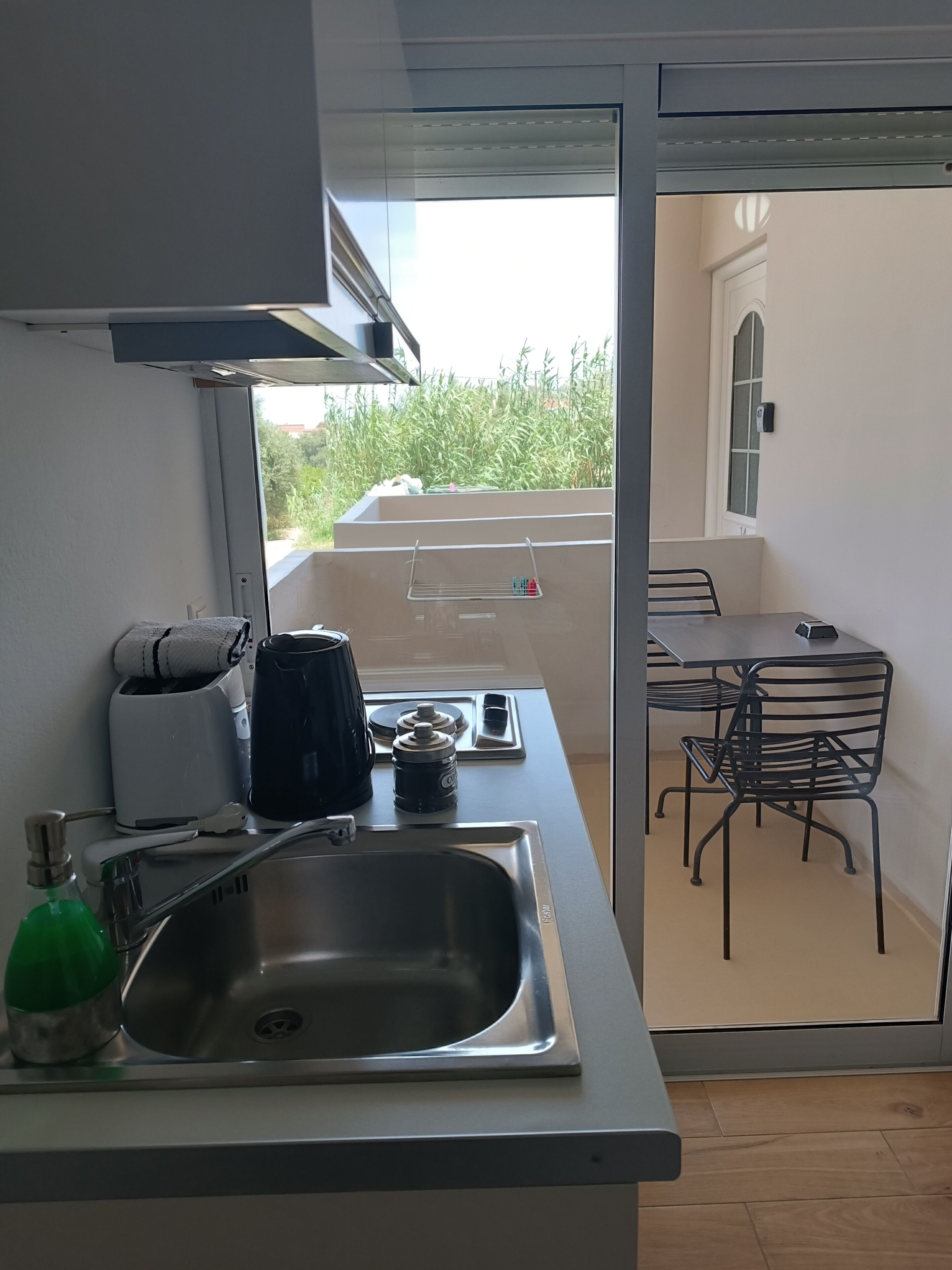 A modern kitchen with a sink, a coffee maker, and a kettle. There is a small table with chairs and a view of greenery outside.