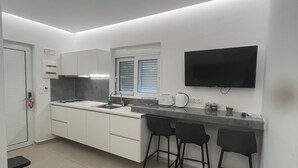 A modern kitchen with white cabinetry, a built-in oven, a microwave, a kettle, and a flat-screen TV mounted on the wall. - Aestas Apts (Chania)