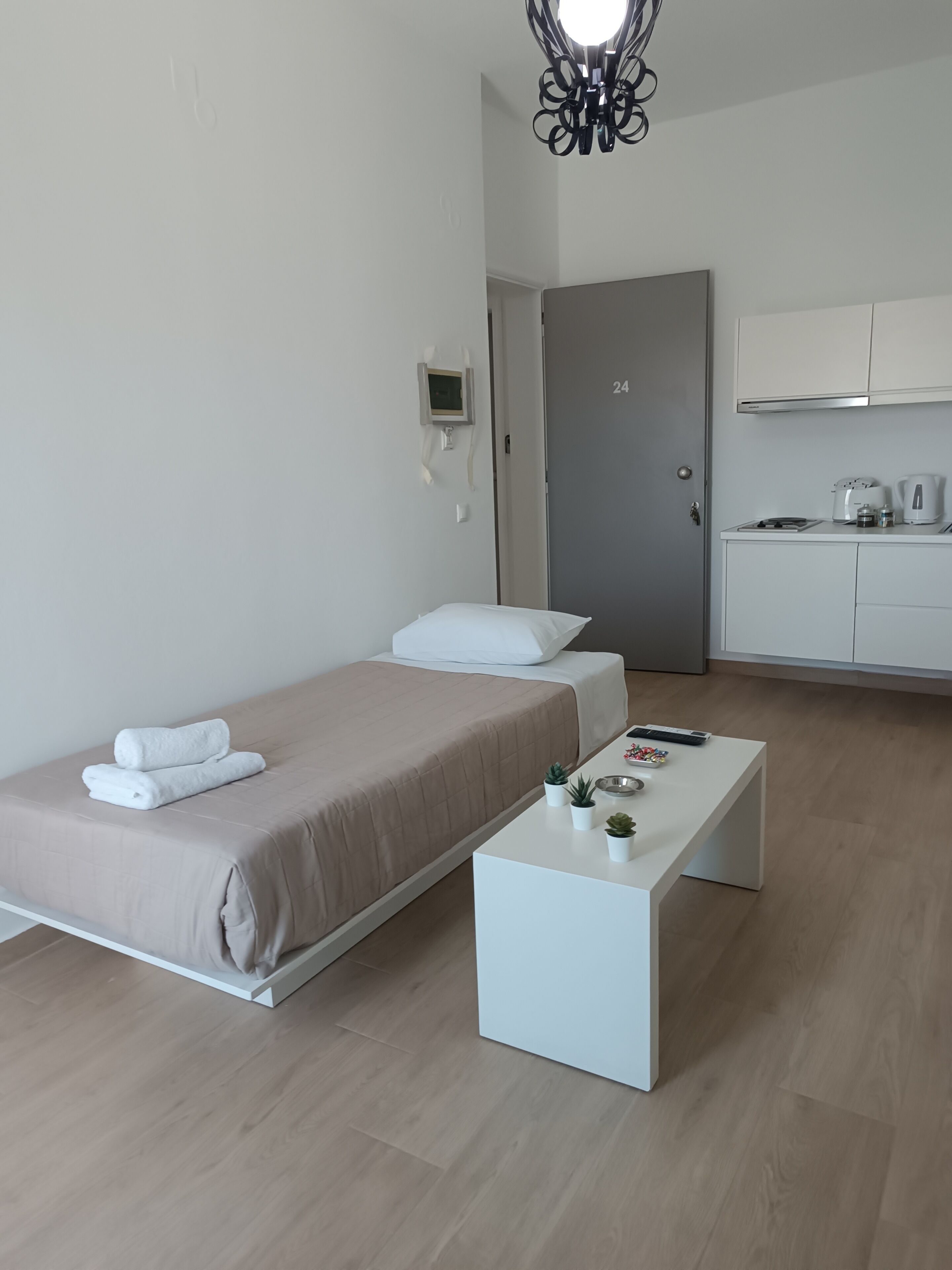 A modern hotel room with a bed, bedside table, and a kitchenette area.