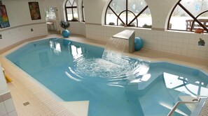 An indoor swimming pool with a water feature, tiled walls, and windows with decorative elements. - Landhotel Basler Hof (Lauterbach)