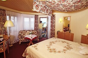 A hotel room with a large bed, a floral-patterned sofa, a small table with a vase, and a wooden desk. - Landhotel Basler Hof (Lauterbach)