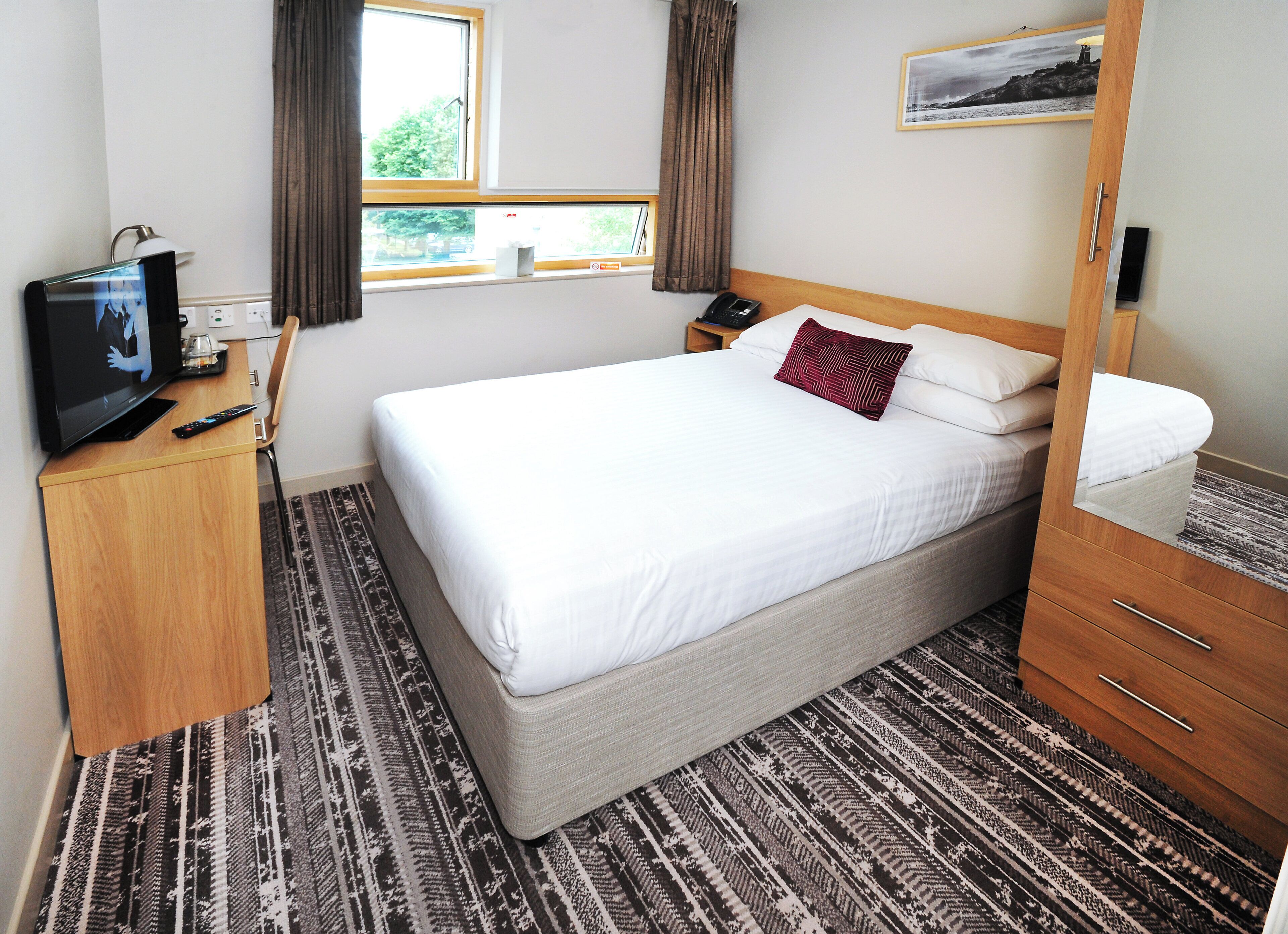 A hotel room with a large bed, a TV on a wooden desk, a bedside table with a lamp, and a window with curtains.
