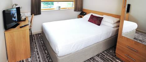 A hotel room with a large bed, a TV on a wooden desk, a bedside table with a lamp, and a window with curtains.