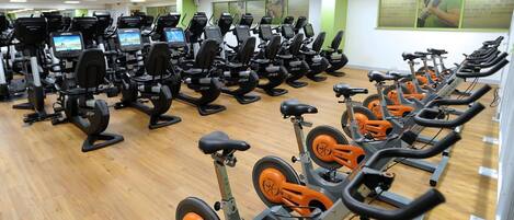 Un gimnasio con filas de bicicletas estáticas y cintas de correr, suelo de madera y paredes verdes.