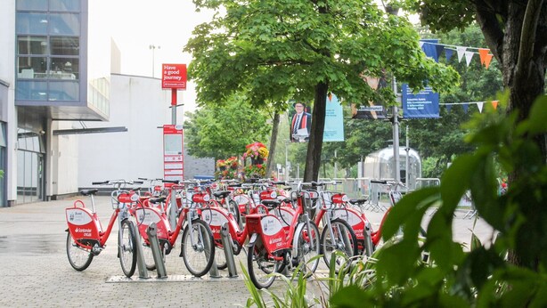 Uma fileira de bicicletas vermelhas Santander estacionadas em área externa com calçada de tijolos, cercada por vegetação e edifícios ao fundo.