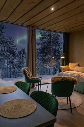 A cozy room with a large window, a green sofa, a green chair, and a table with woven placemats. - Tankavaara Gold Village (Sodankyla)