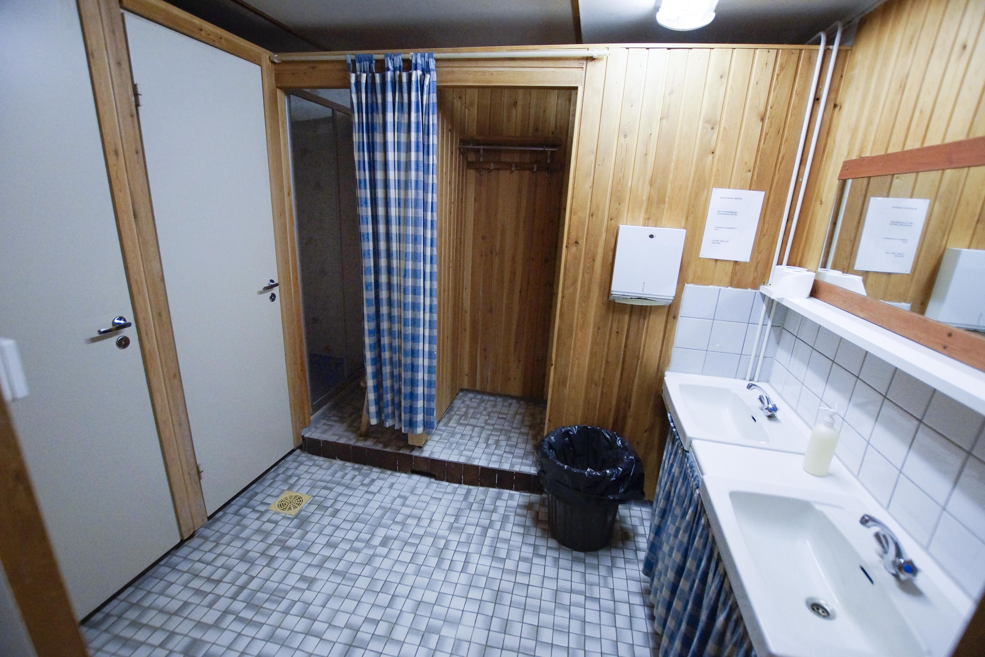 A bathroom with a wooden shower enclosure, a tiled floor, a sink with a mirror, and a trash bin.