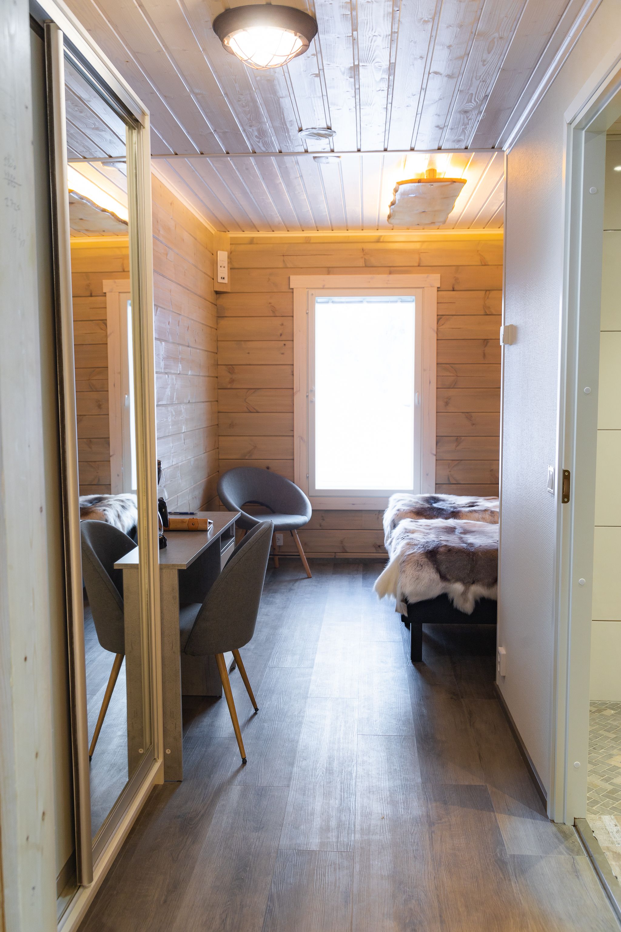 Ein gemütlicher Raum mit Holzboden, einem Schreibtisch mit Stuhl, einem Bett mit Felldecke und einem großen Fenster, das viel Tageslicht hereinlässt.