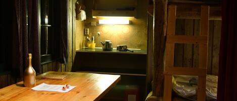 Una mesa de madera con una botella y papeles, un banco, una litera de madera y una cocina con una lámpara sobre la encimera.