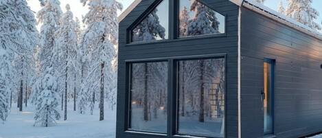 A modern cabin with a large glass window and a sloped roof, surrounded by snow-covered trees.