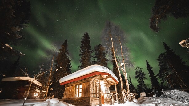 A wooden cabin with a snow-covered roof, surrounded by snow and trees, under a starry night sky with the aurora borealis. - Tankavaara Gold Village (Sodankyla)